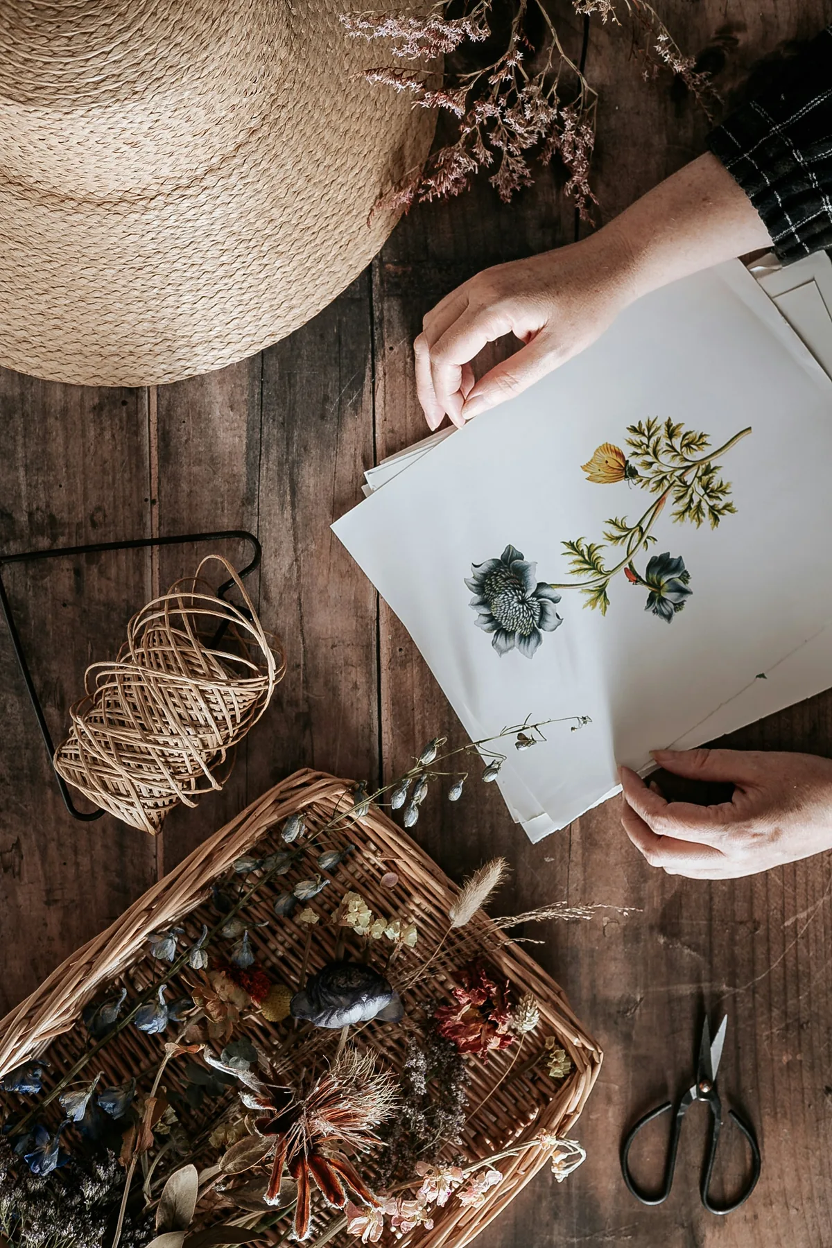 Manos preparando una composición botánica con flores secas sobre una mesa de madera